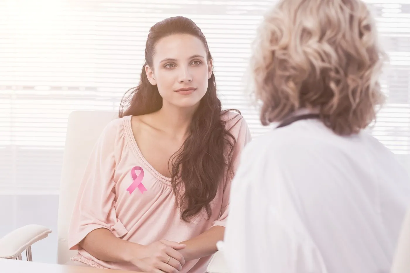 Jovem em consulta médica com símbolo rosa na blusa, indicando cuidados e prevenção contra câncer de mama em mulheres mais jovens.