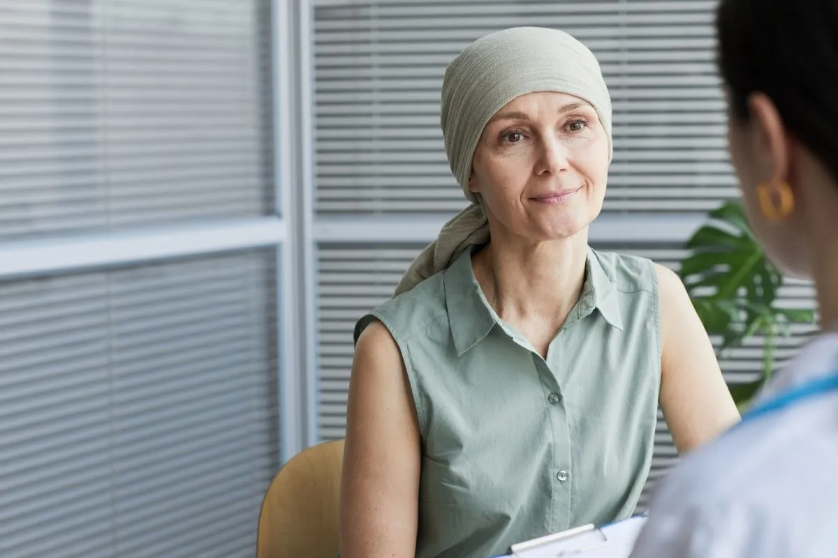 Mulher com câncer de mama com metástase na axila tem cura com lenço na cabeça conversando com mastologista durante consulta.